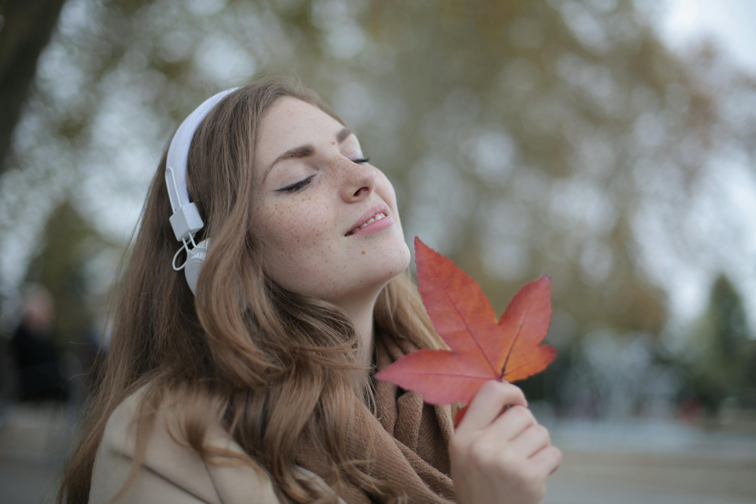 Young woman with headphones holding a red leaf, enjoying music outdoors in autumn.